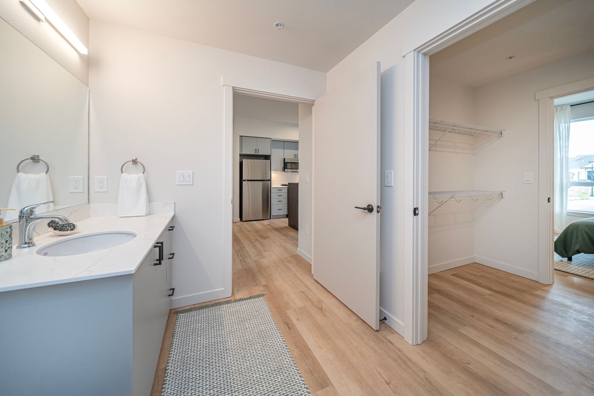 Bathroom with vanity, walk-in closet, and doorway to kitchen. Light wood floors, white walls, and gray cabinets.