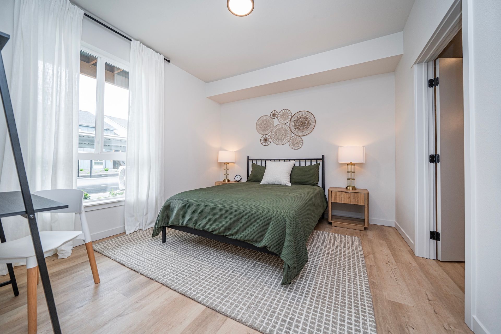 Bedroom with a green bedspread, wooden floors, and a desk by a large window with white curtains.
