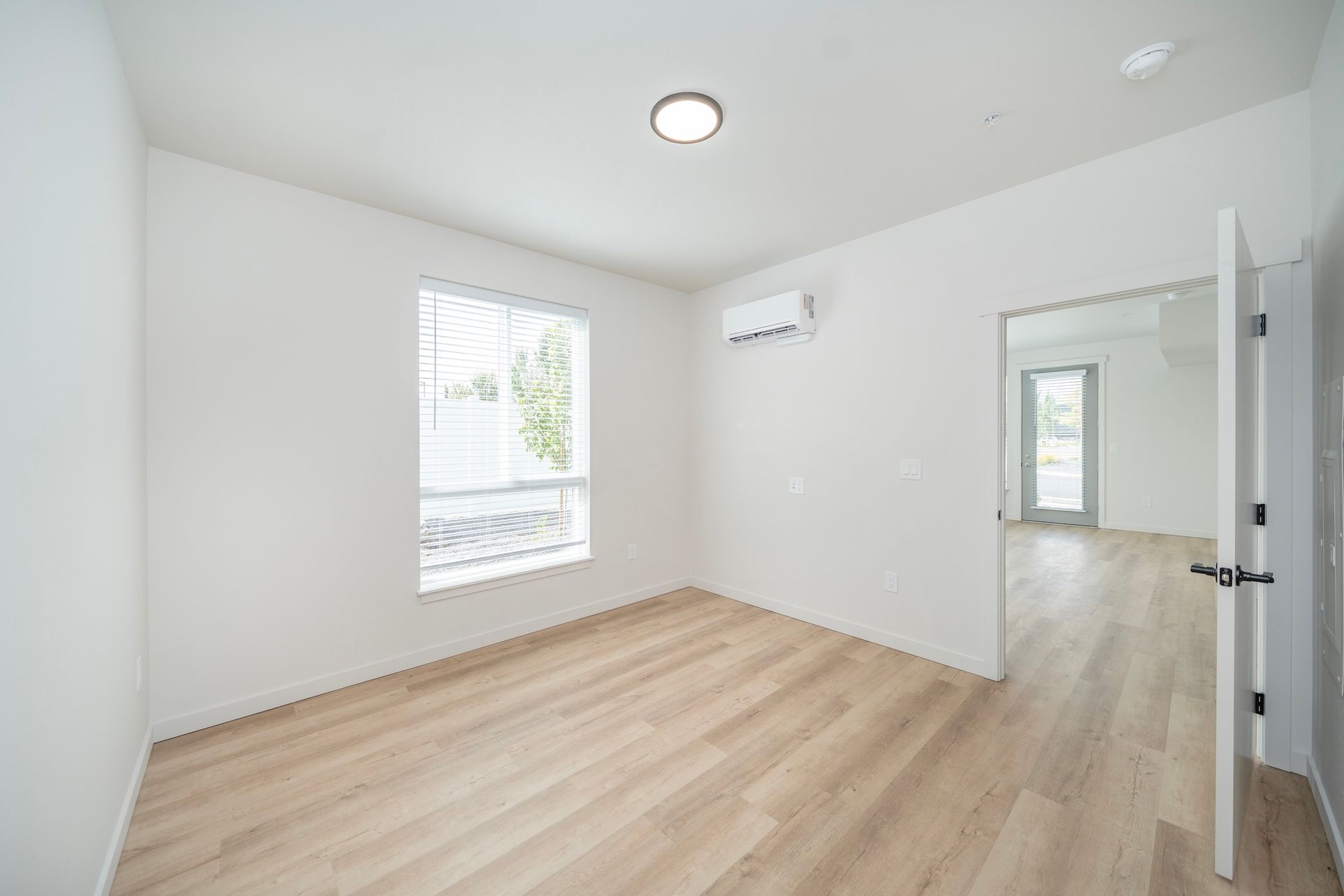Empty room with light wood floors, a window, and an open doorway; walls and ceiling are white.