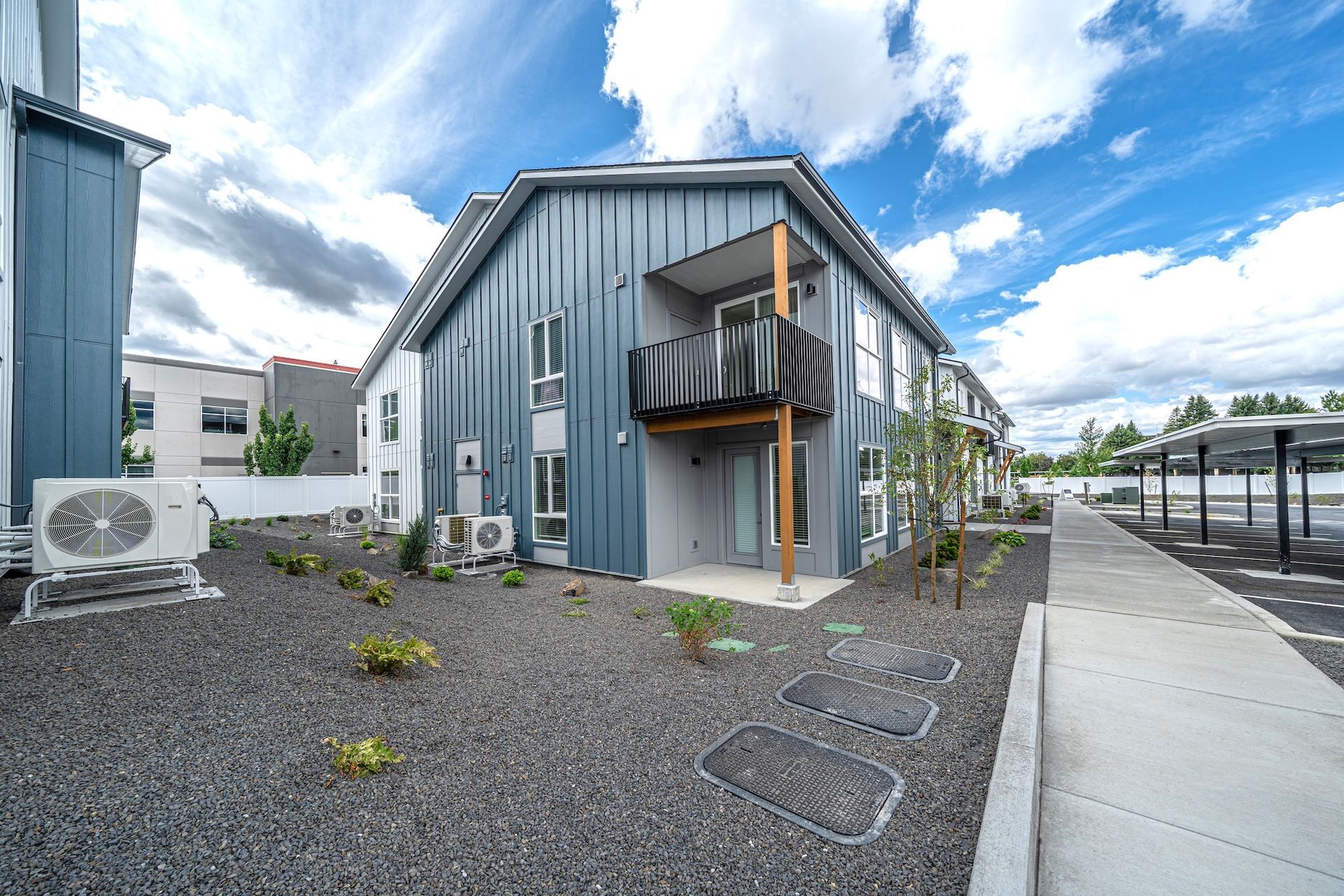 Two-story blue apartment building with a balcony, gravel yard, and sidewalk.