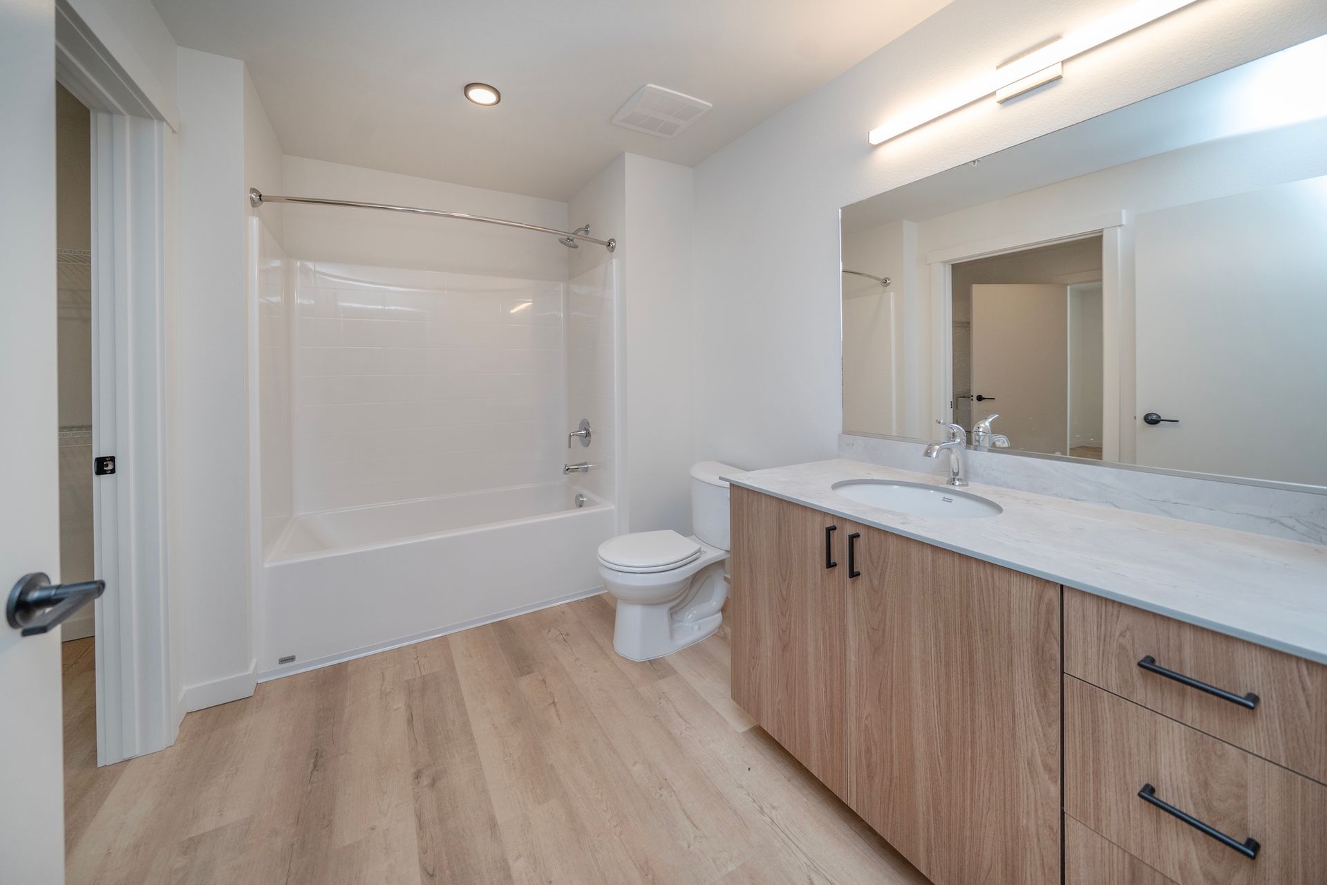 Bathroom with a white tub, toilet, and sink with a wood vanity. Light wood flooring and white walls.