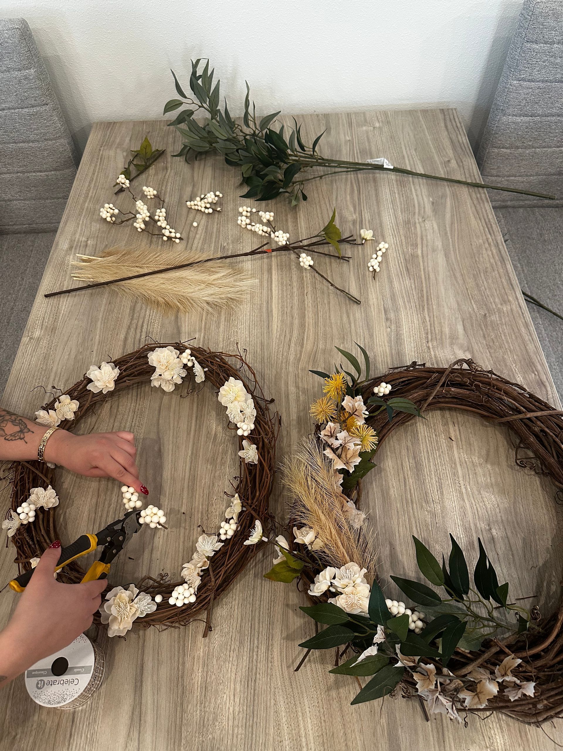 Hands using wire cutters to attach white floral sprigs to a twig wreath on a table with more greenery nearby.