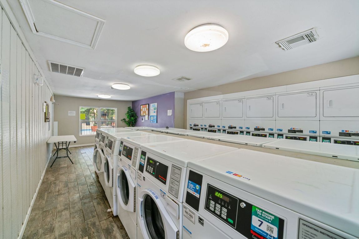 A laundromat with many washing machines and a folding table. Light-colored machines on a wood-look floor, purple accent wall.