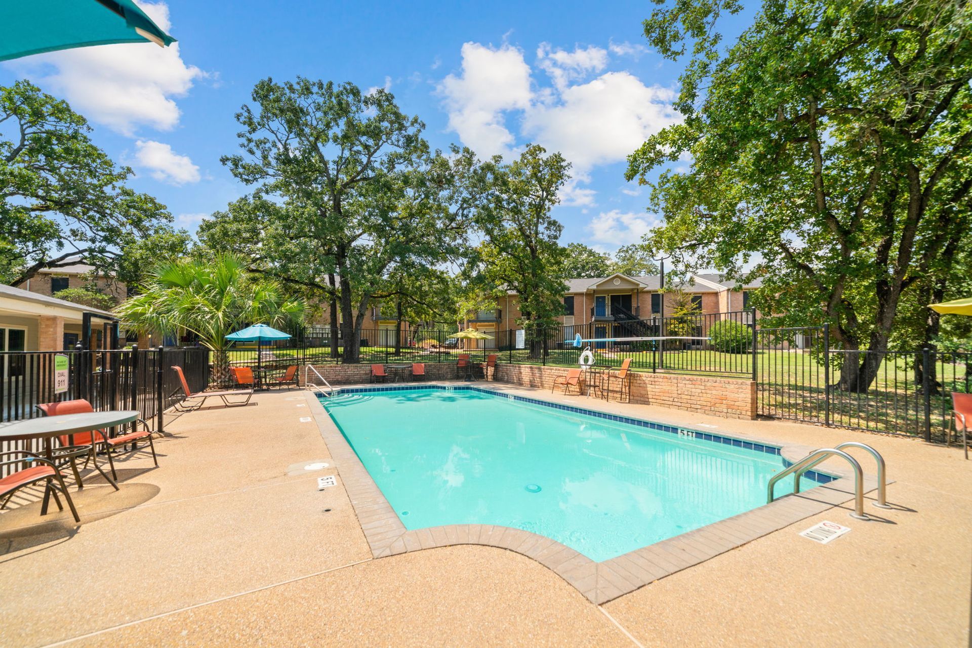 Pool area with blue water, surrounded by concrete, trees, and small buildings.