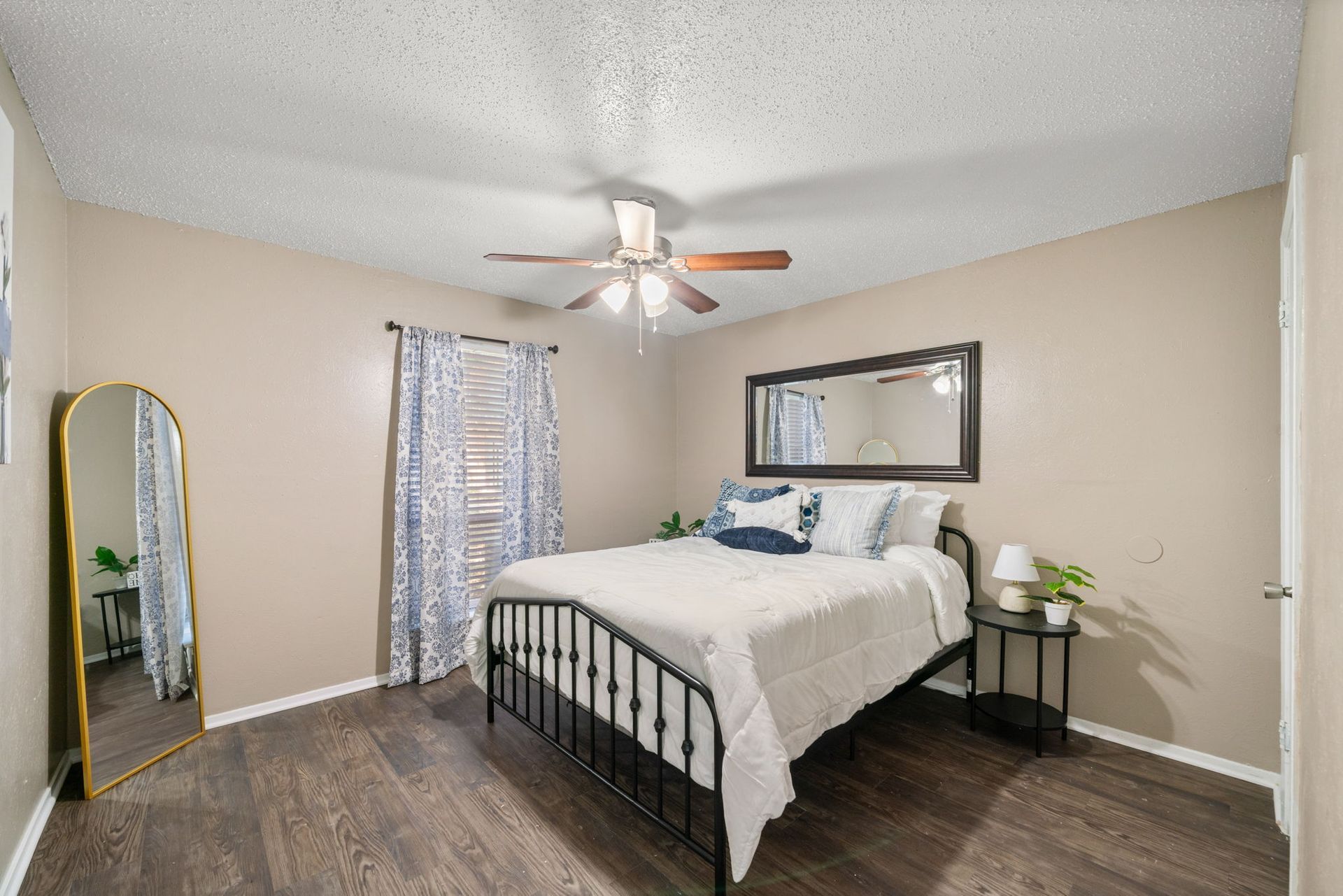 Bedroom with a black bed, mirror, window with blue curtains, and dark wood flooring.