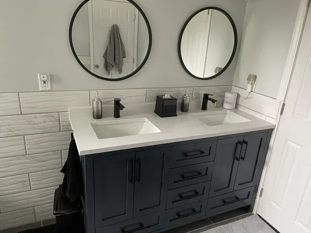 Bathroom vanity with blue cabinets, white countertop, two sinks, black faucets, mirrors, and white tiled wall.
