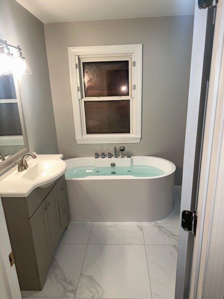 Gray bathroom with an oval tub filled with water, vanity, window, and tile floor.