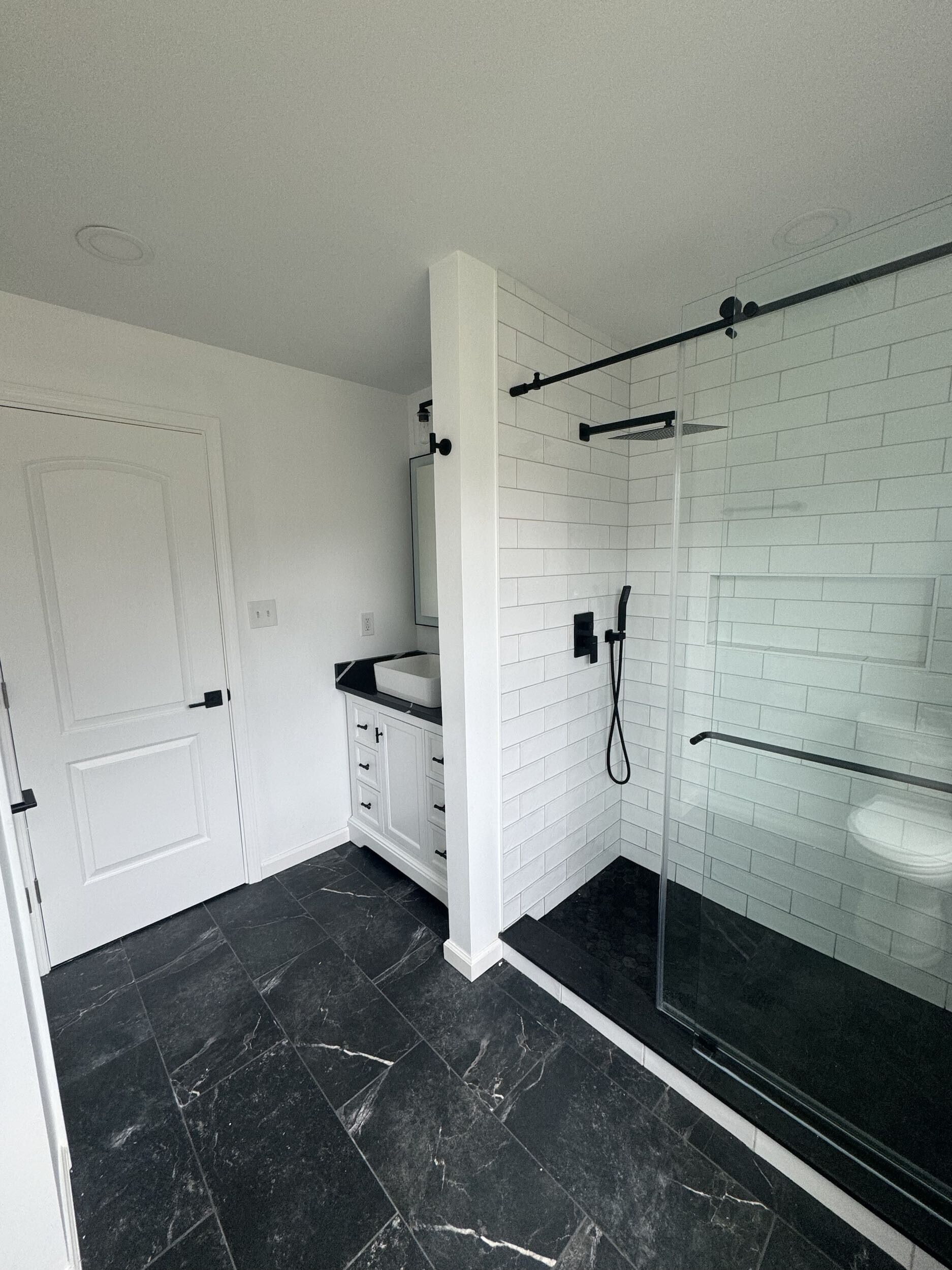 Bathroom with black marble floor, white tiled shower, black hardware and white vanity.