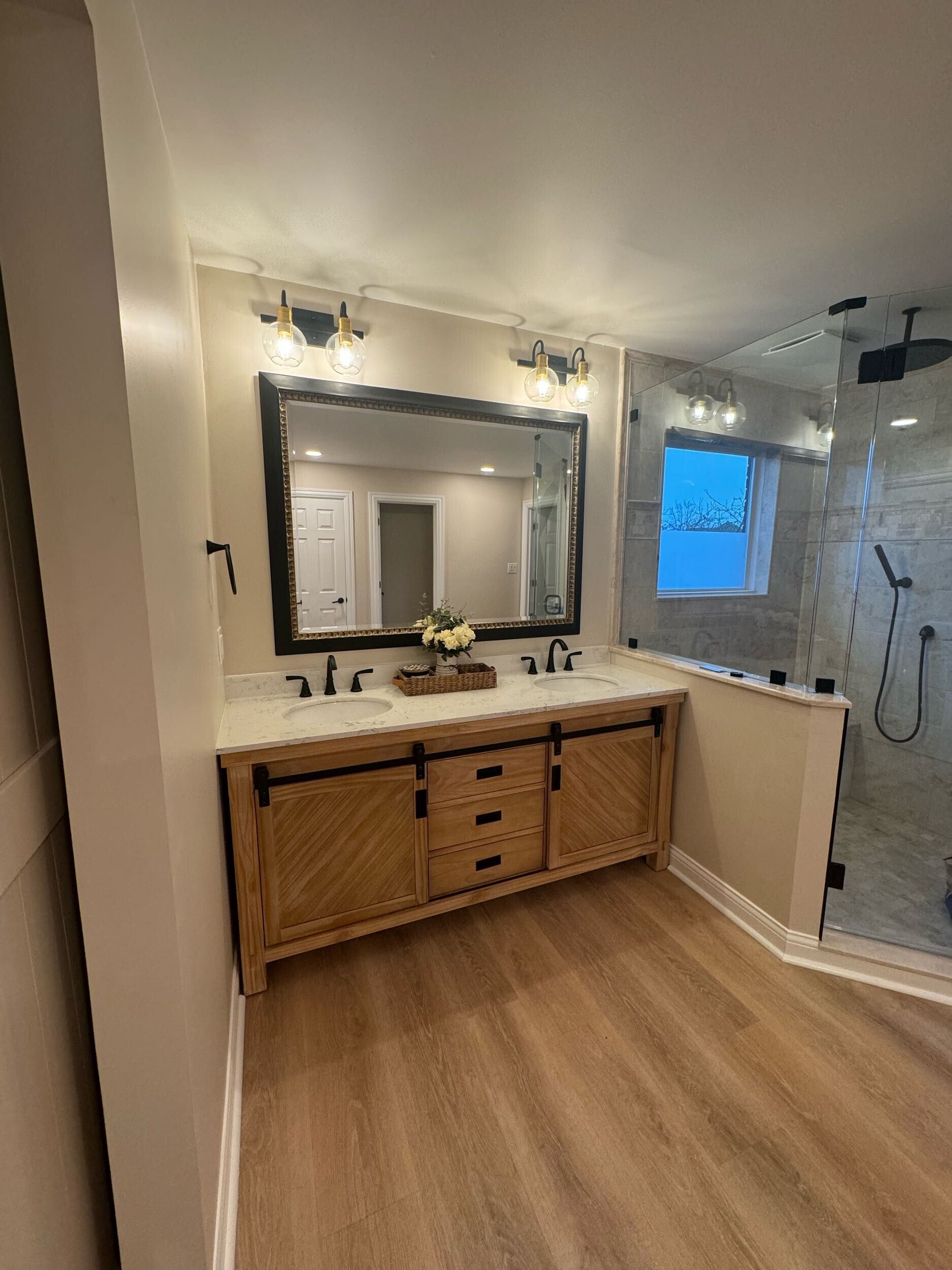 Modern bathroom with wood vanity, large mirror, and glass shower, lit by wall sconces and natural light.