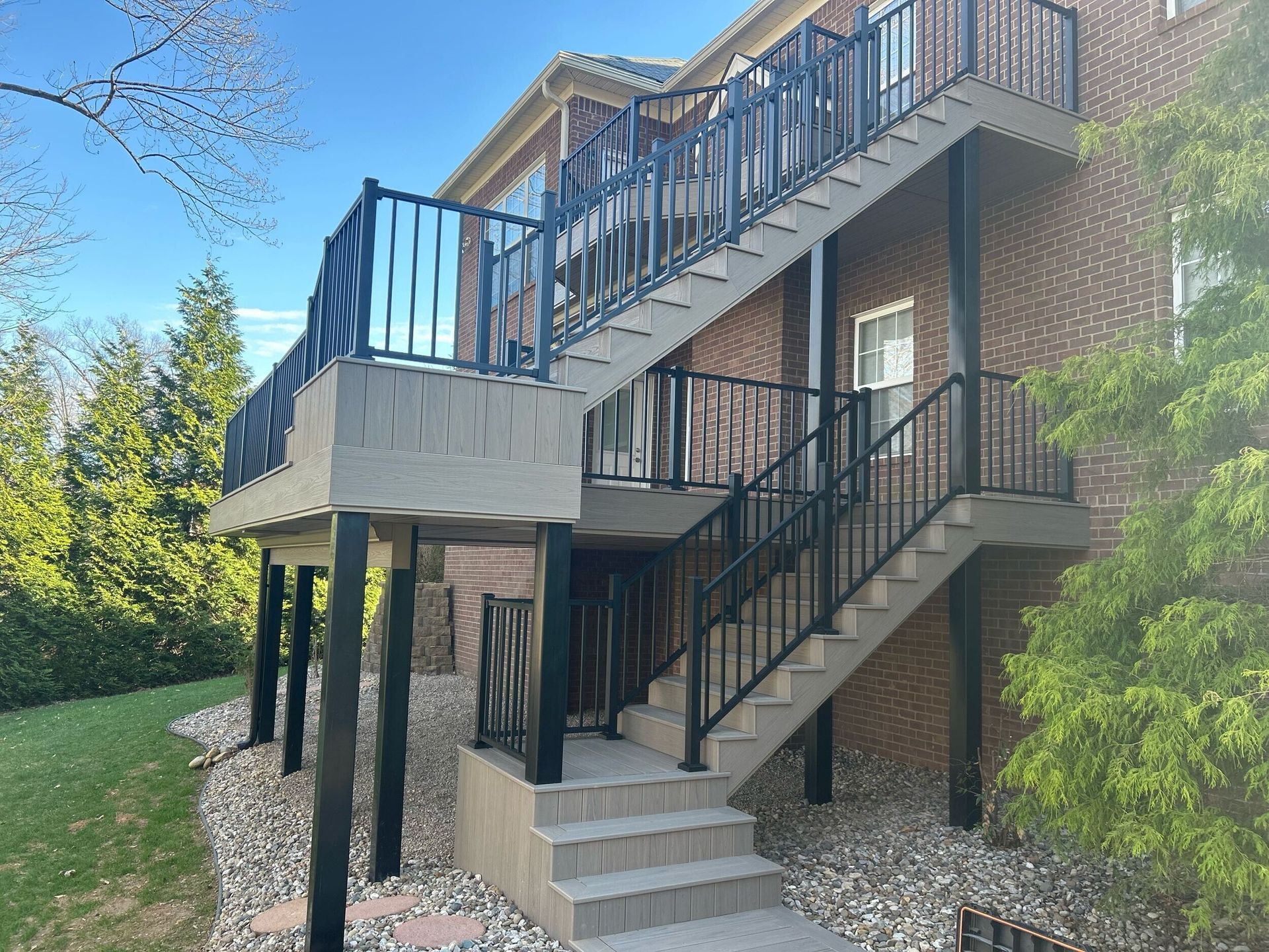 Multi-level deck and stairs with black railings attached to a brick building, with greenery and blue sky.