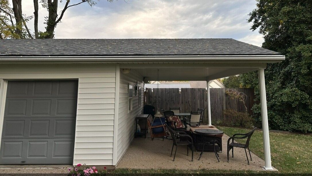 Garage with attached covered patio; gray door, white siding, patio furniture, backyard setting.