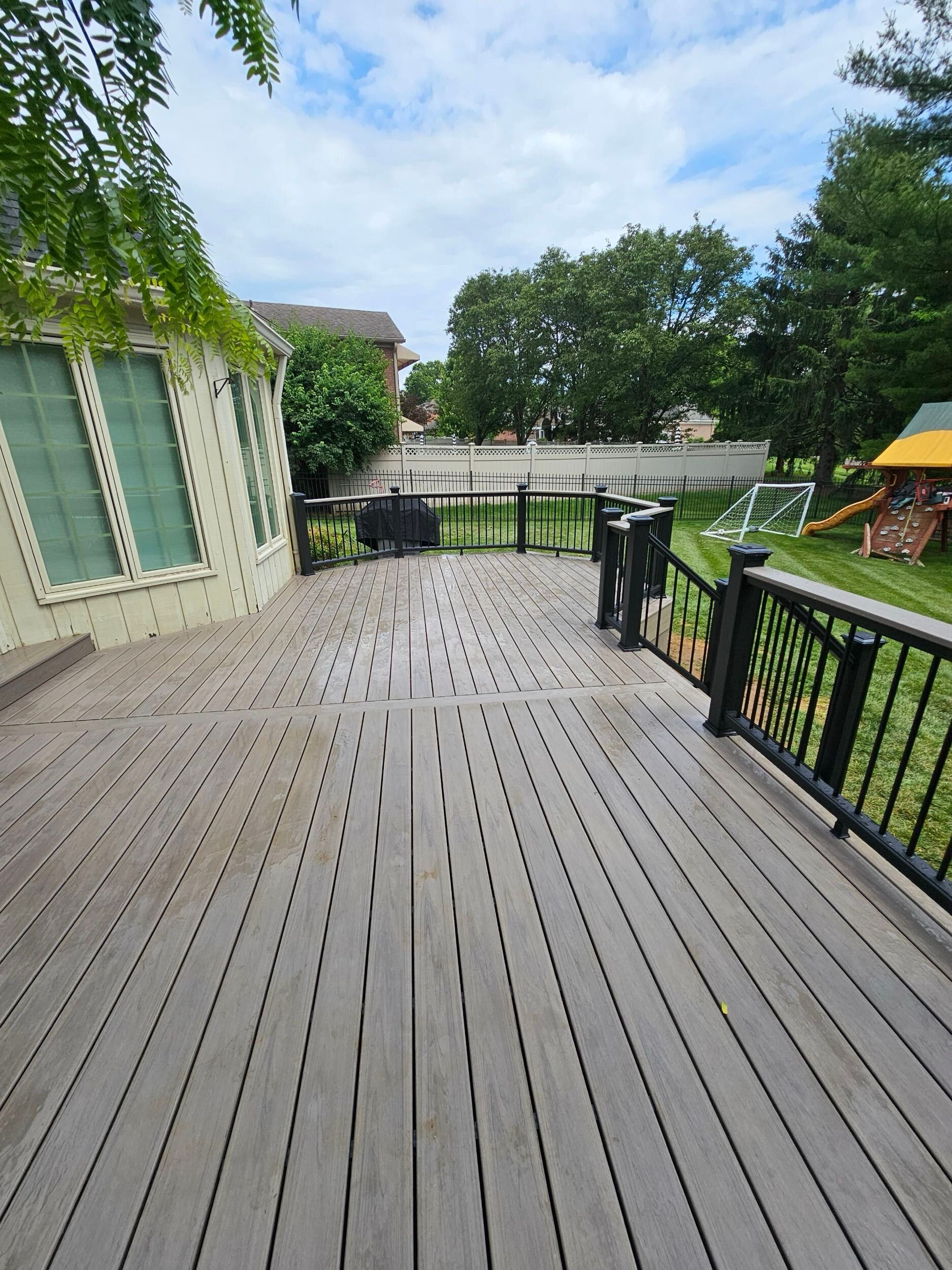 Wooden deck with dark railing overlooks a backyard with trees, a swing set, and a soccer net.