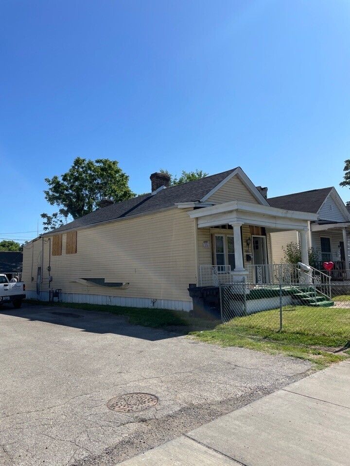 Single-story beige house with porch, chain-link fence, and driveway under a clear blue sky