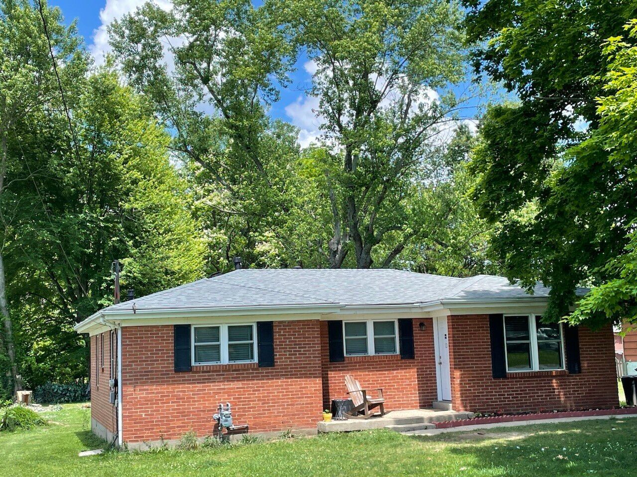 Red brick ranch house with black shutters, under a cloudy blue sky, surrounded by green trees.