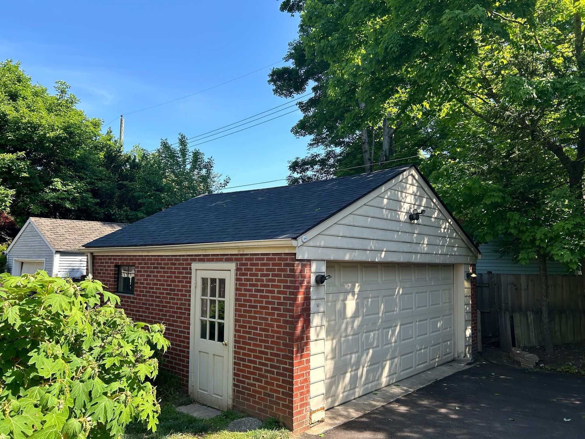 Small brick garage with white door and gray shingled roof, surrounded by trees on a sunny day