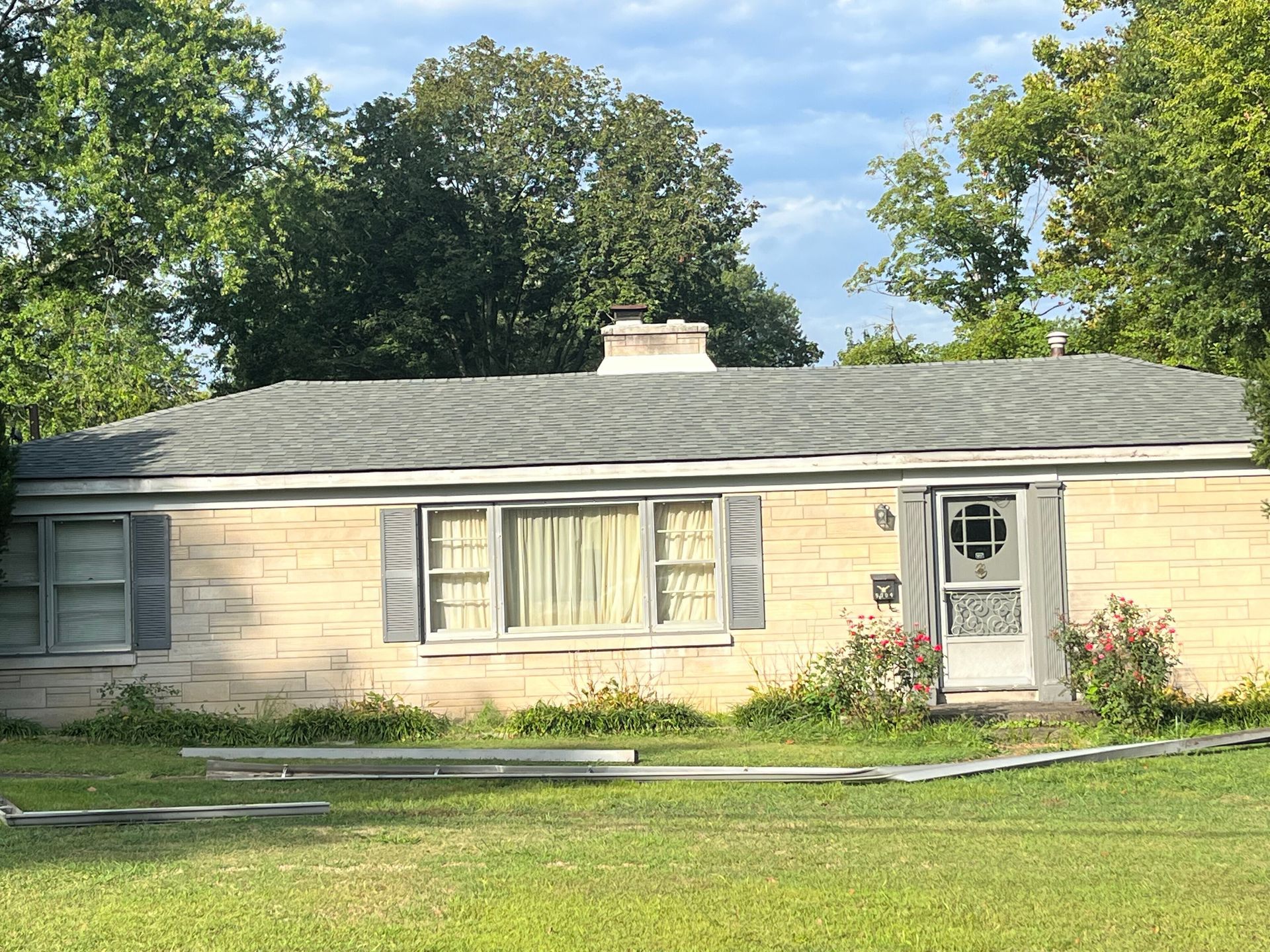 Single-story house with pale brick walls, gray roof, and overgrown yard. Door has a decorative screen.