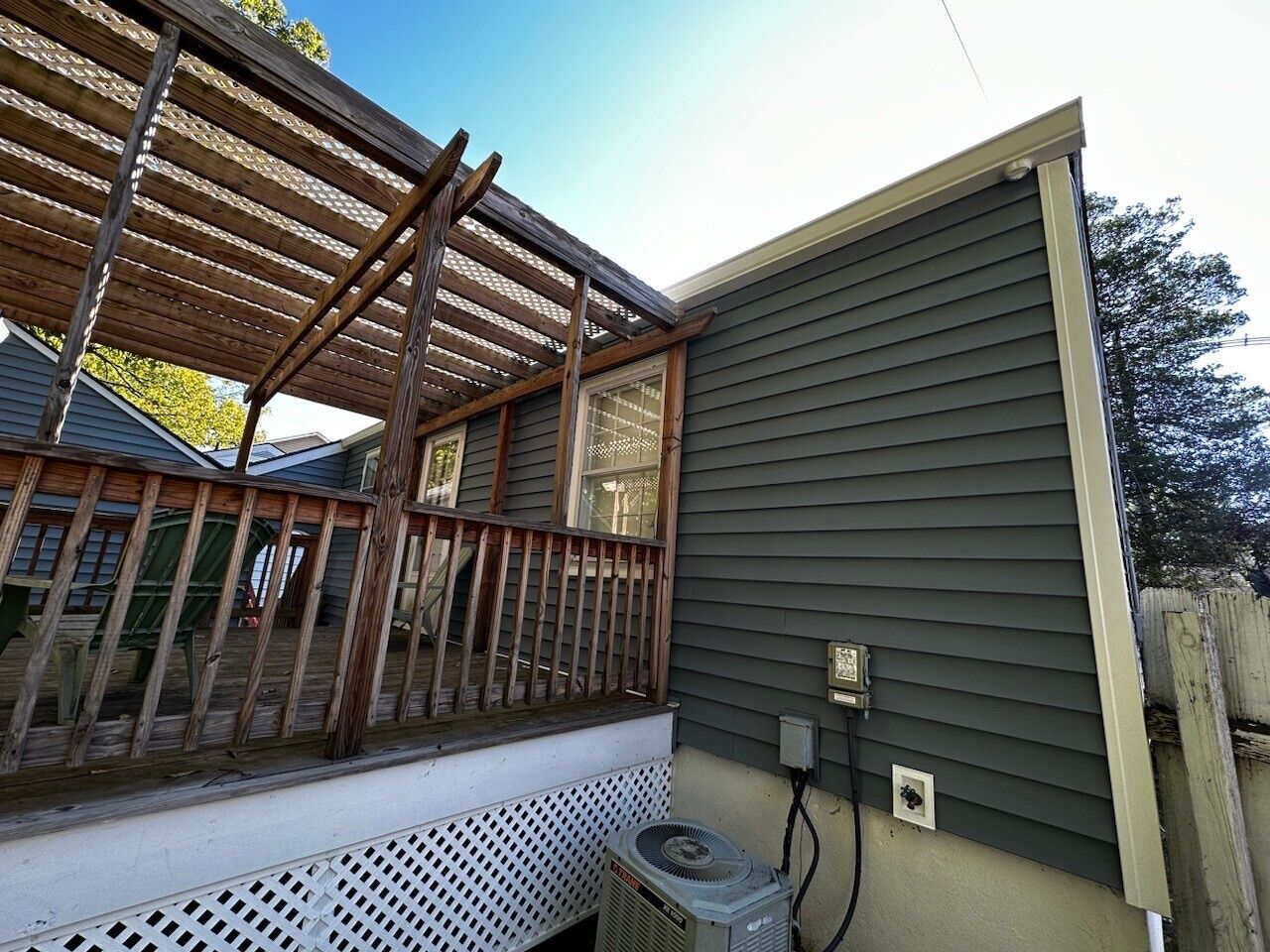 Back deck with pergola, wooden railing, and green house siding with utility outlets.