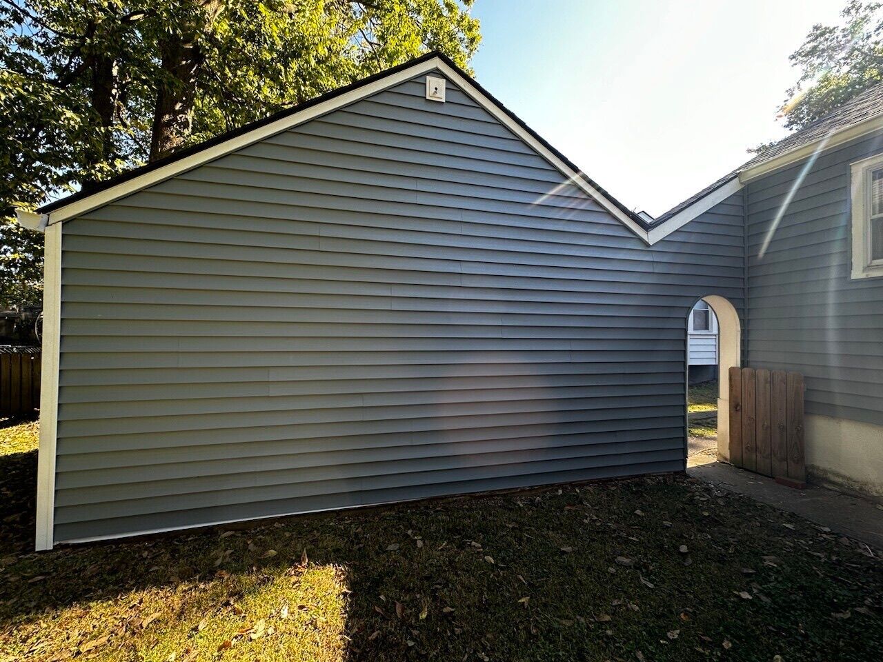 Gray vinyl-sided house exterior with gabled roof and small arched doorway in a sunlit yard
