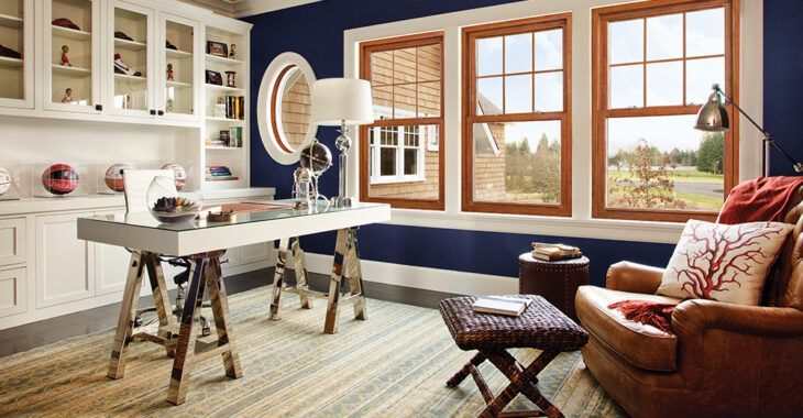 A living room with blue walls , white cabinets , a desk , a chair , and two windows.