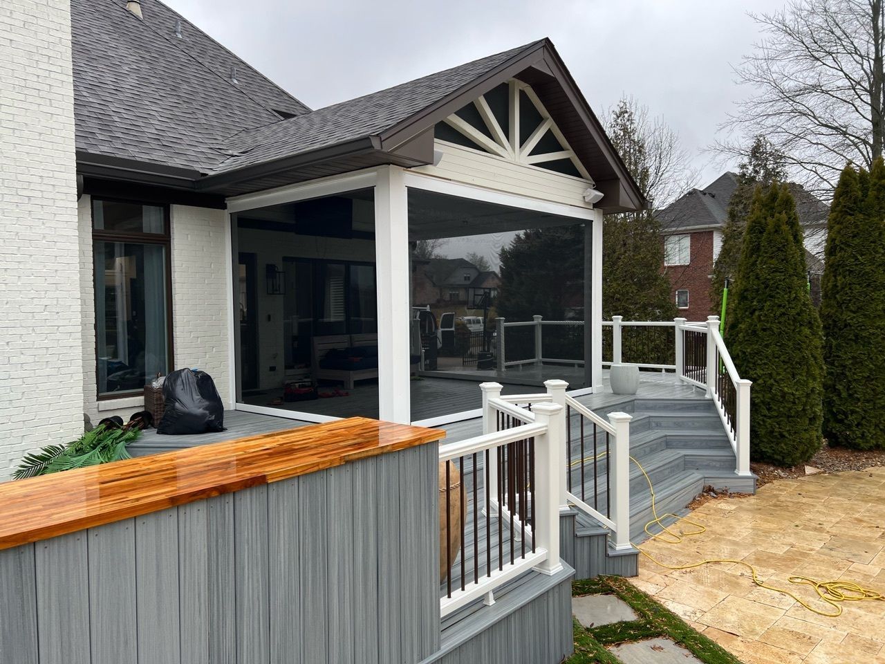 Screened-in porch with white trim, gray deck, and brown roof.  Tall evergreens and a brick building in the background.