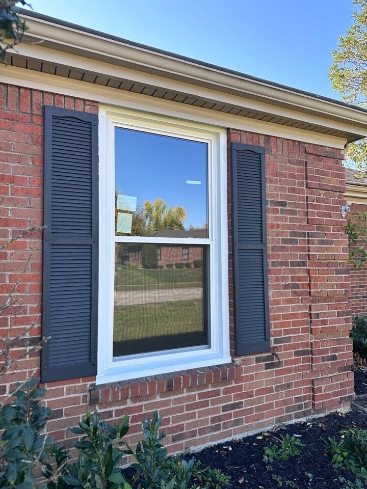 Brick house window with white trim and black shutters beside a landscaped bed