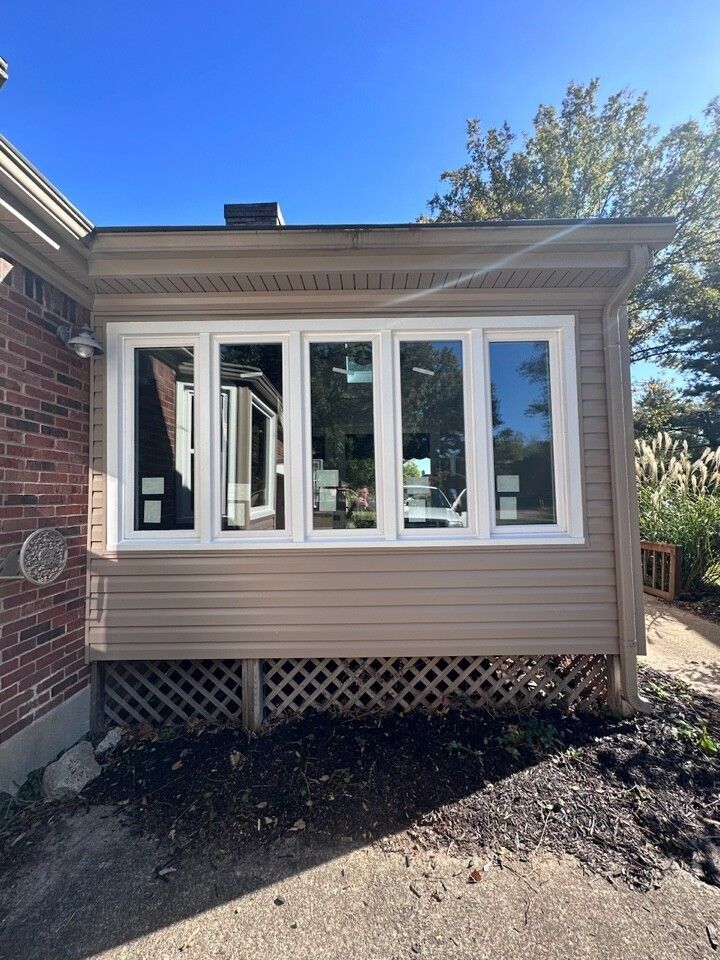 Enclosed porch with white-framed windows and tan siding on a brick house under a clear blue sky