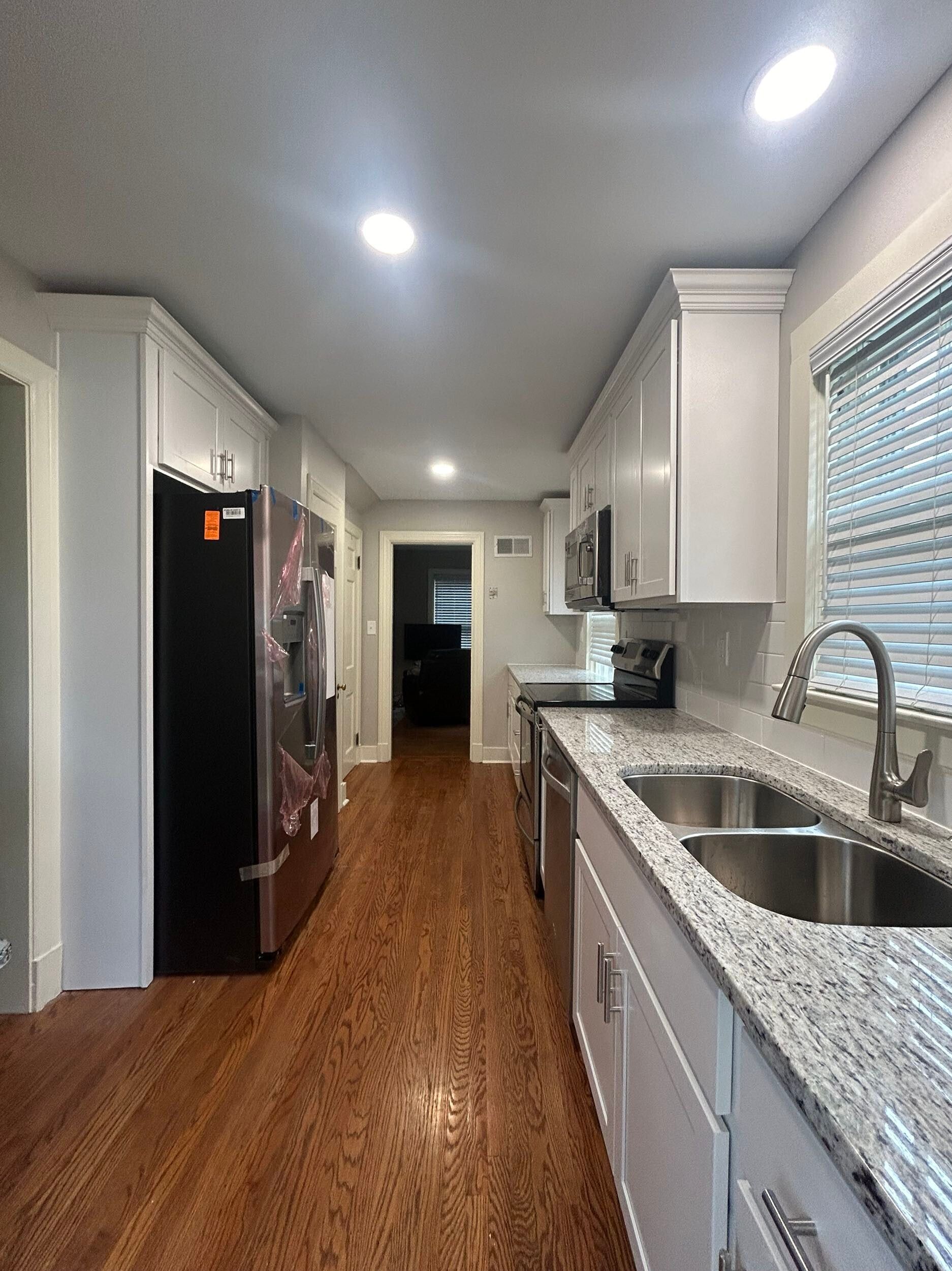 Long, narrow kitchen with white cabinets, stainless steel appliances, and wood flooring.