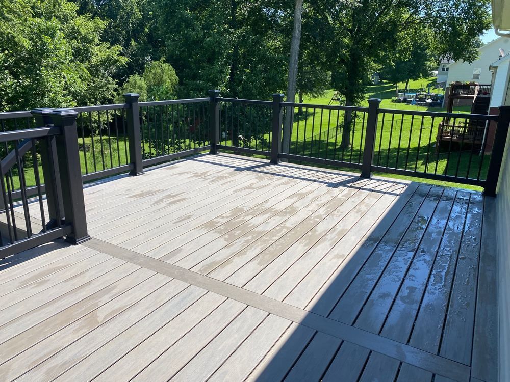 Sunlit wooden deck with black railing, bordered by green trees and lawn