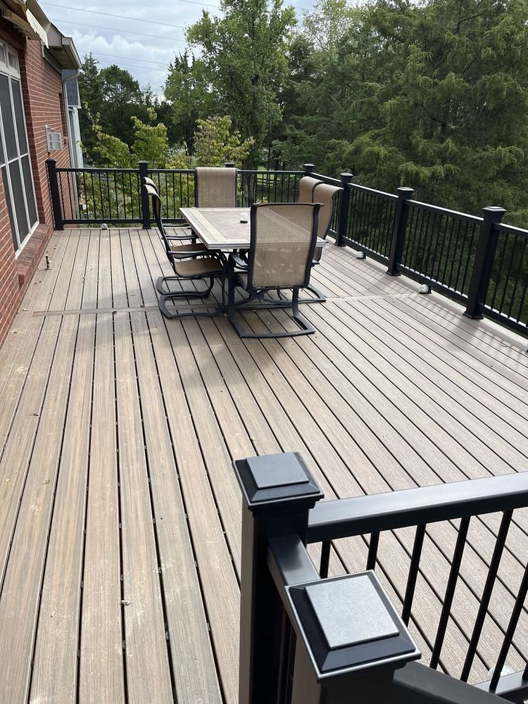 Outdoor wooden deck with black railing, patio table and chairs, and a brick house wall on the left.