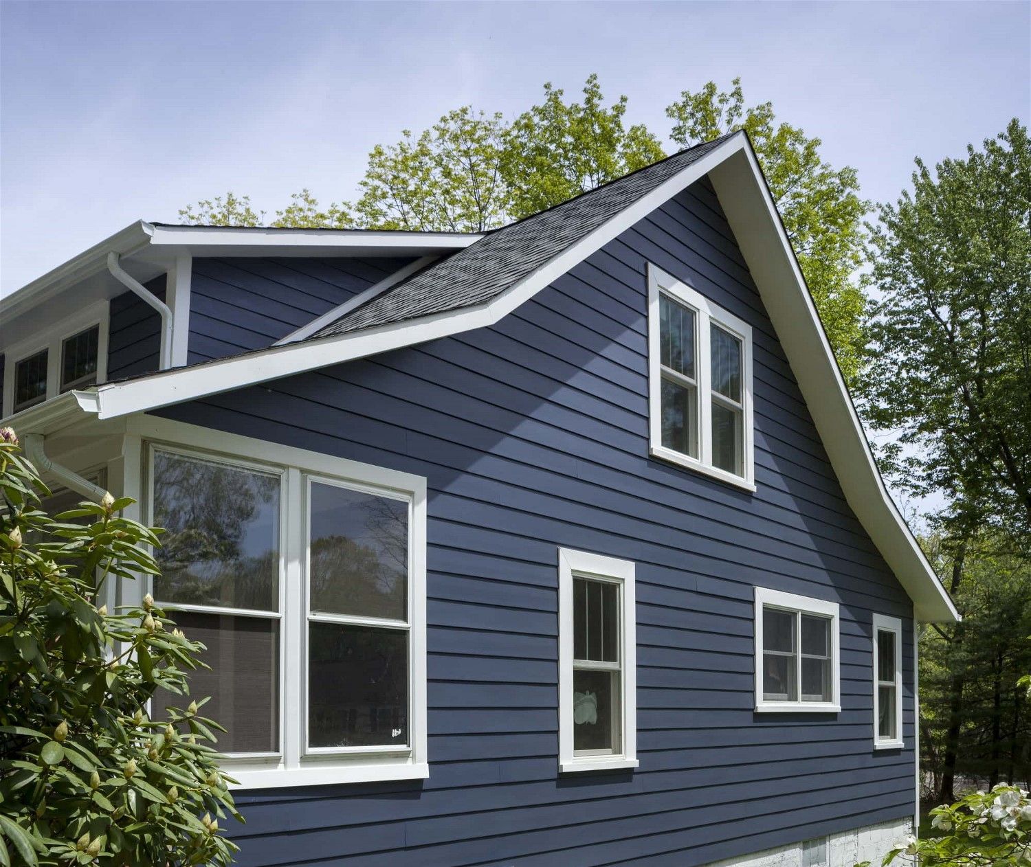 Blue house with white trim and steep roof, surrounded by green trees and shrubs.