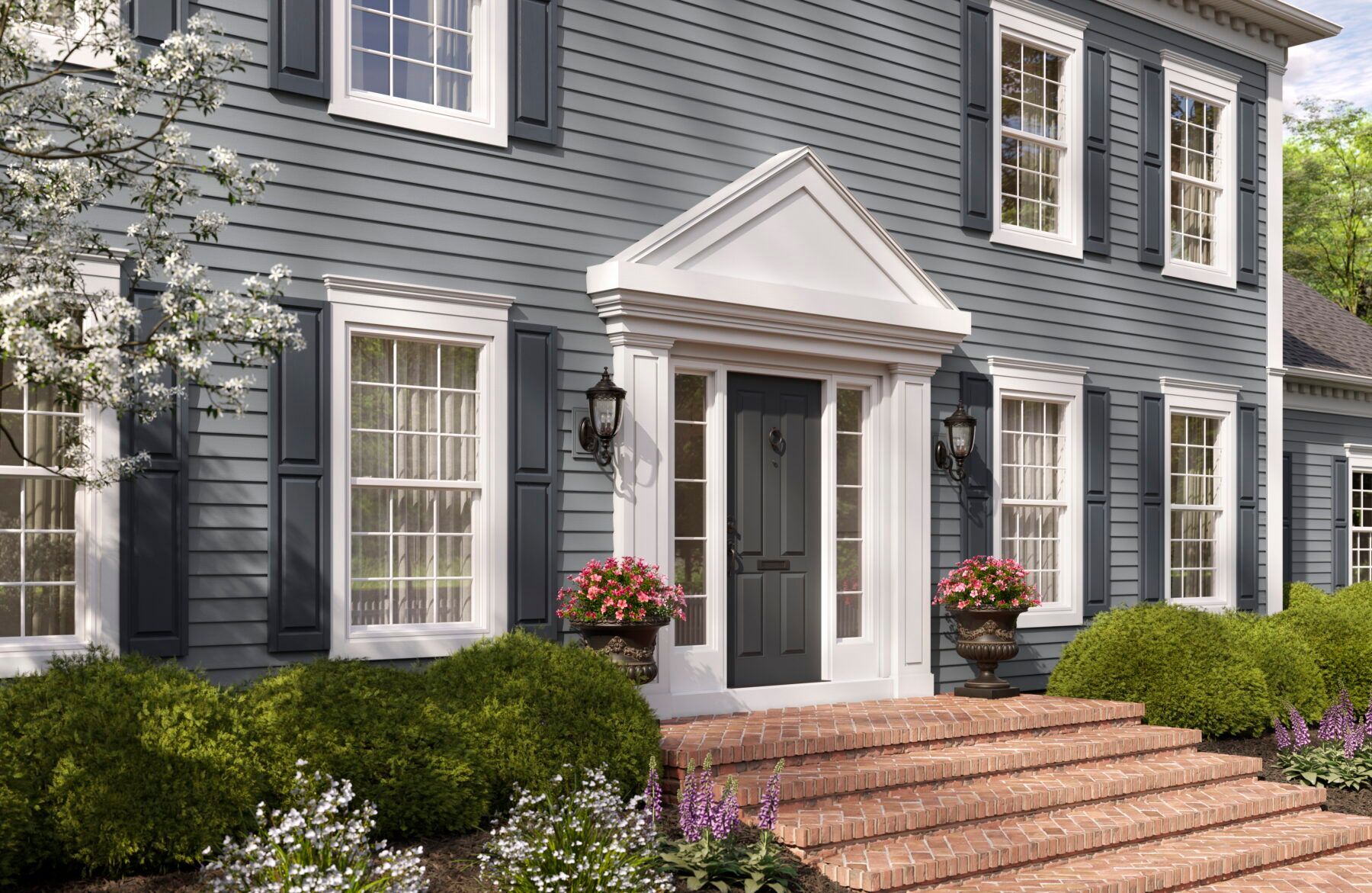 Gray colonial-style house with white trim, black shutters, and a red brick walkway lined by shrubs.