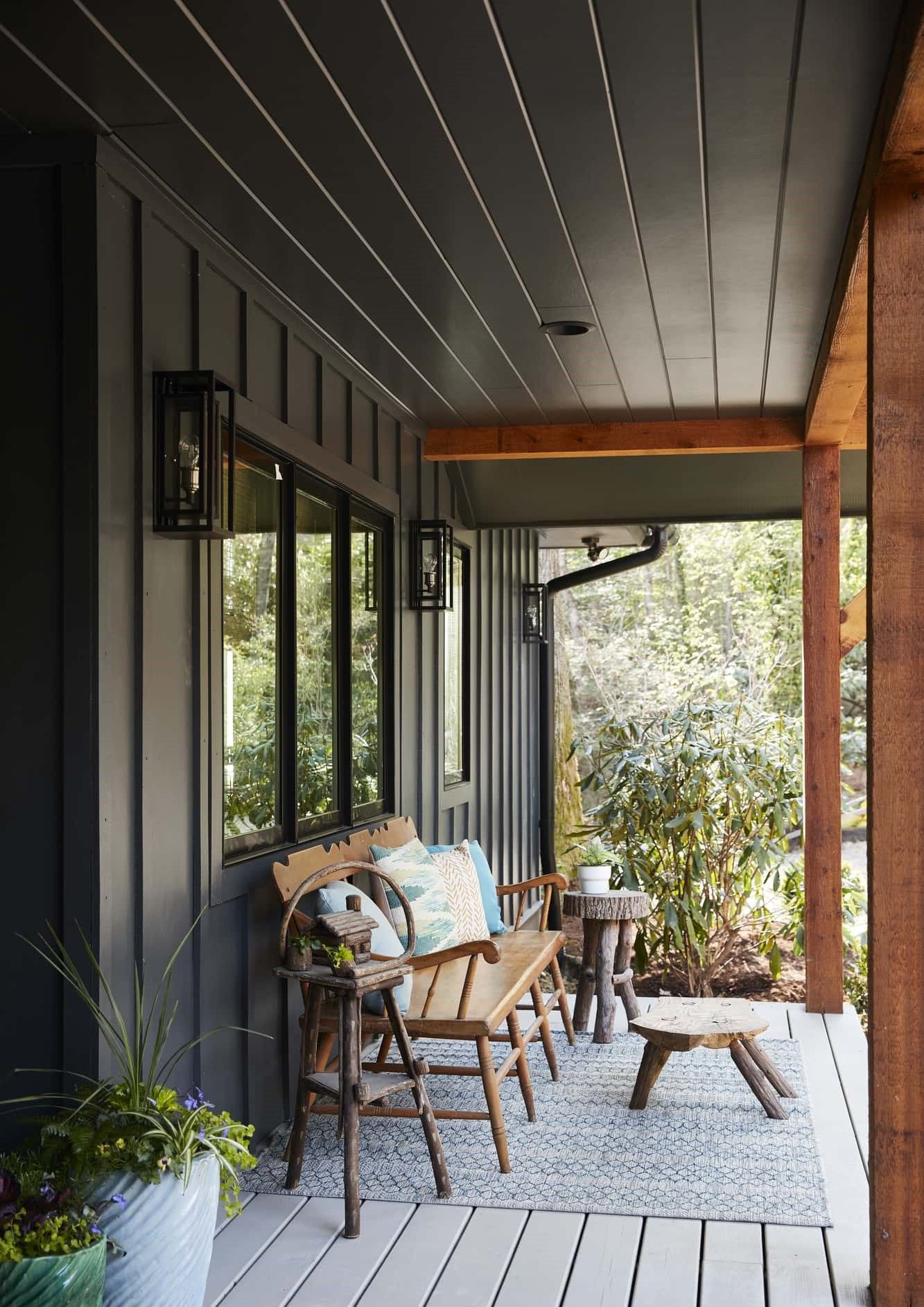 Covered porch with dark siding, wicker chairs, potted plants, and a small table overlooking greenery.