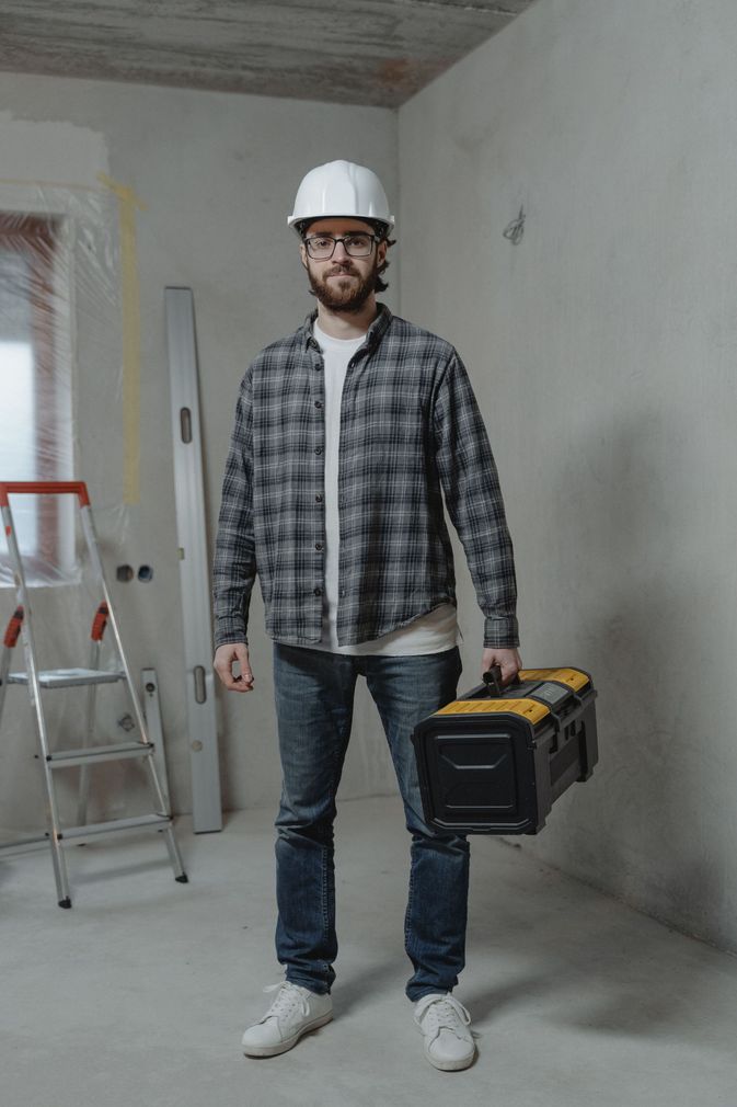Construction worker in hard hat holding a toolbox in an unfinished room with a ladder nearby