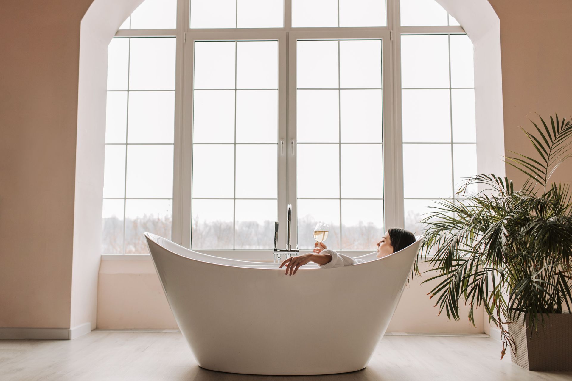 Person relaxing in a white freestanding bathtub by large windows in a bright modern room