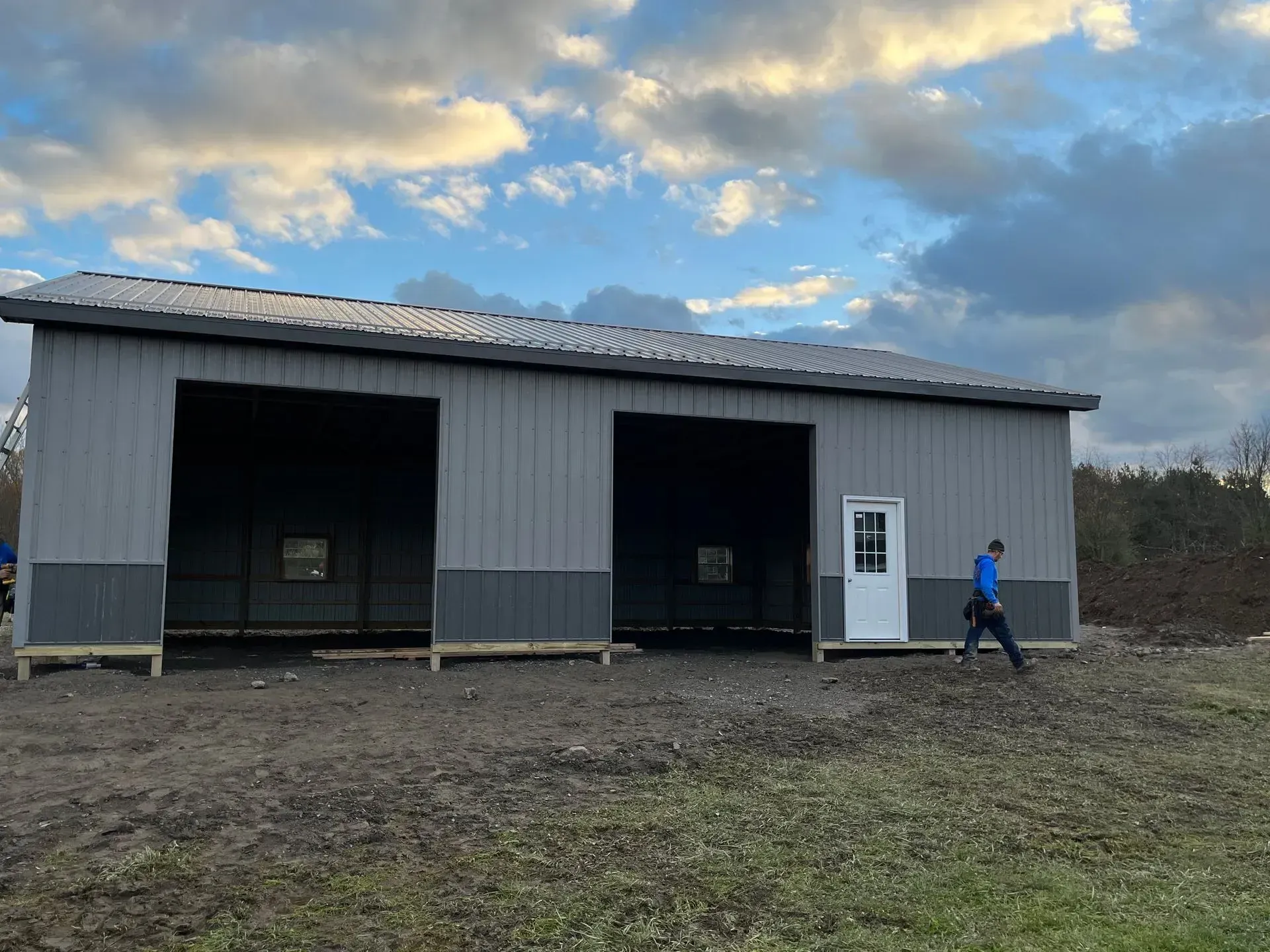 Gray metal building with open bays, person walking toward white door, cloudy sky.