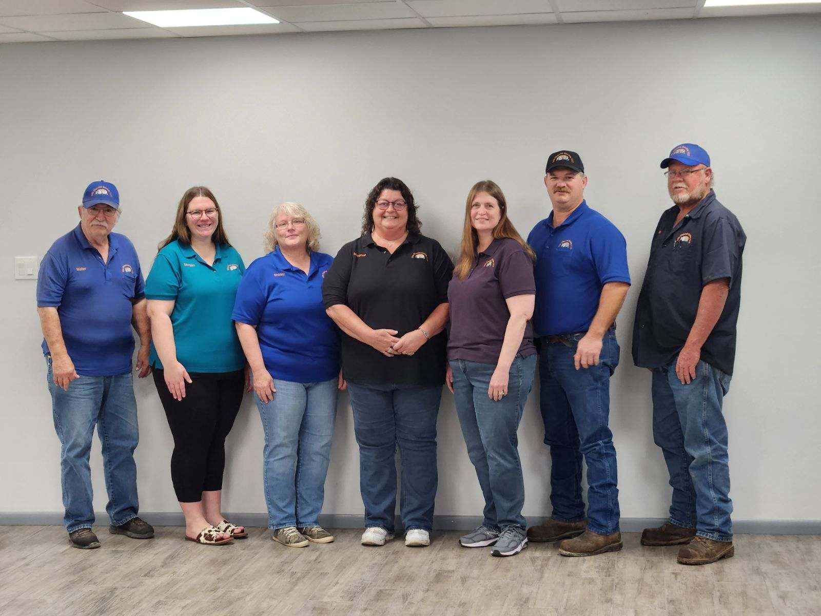 Seven people standing indoors against a plain wall, wearing mostly blue shirts and jeans