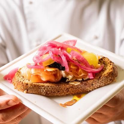 Tramezzino aperto con cipolle sottaceto, salmone affumicato e salsa su un piatto bianco.