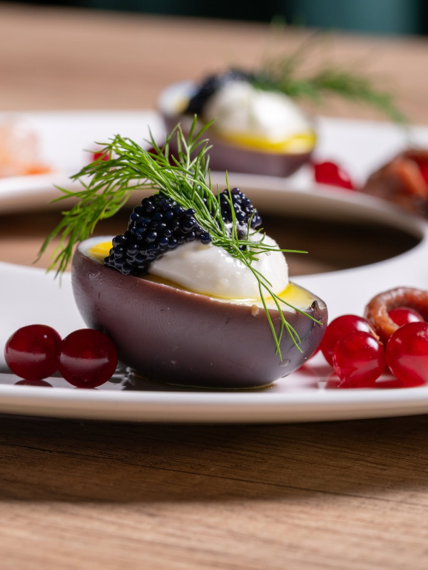 Dessert gourmet in un guscio di cioccolato, guarnito con caviale e aneto su un piatto bianco, con frutti di bosco rossi.