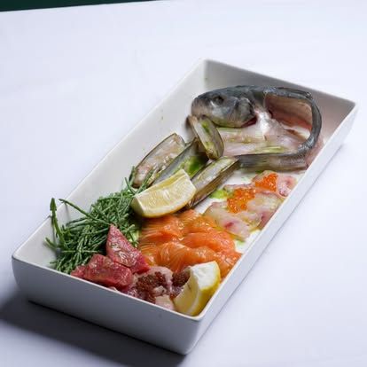 Pesce crudo, spicchi di lime ed erbe aromatiche disposti in un vassoio rettangolare bianco.