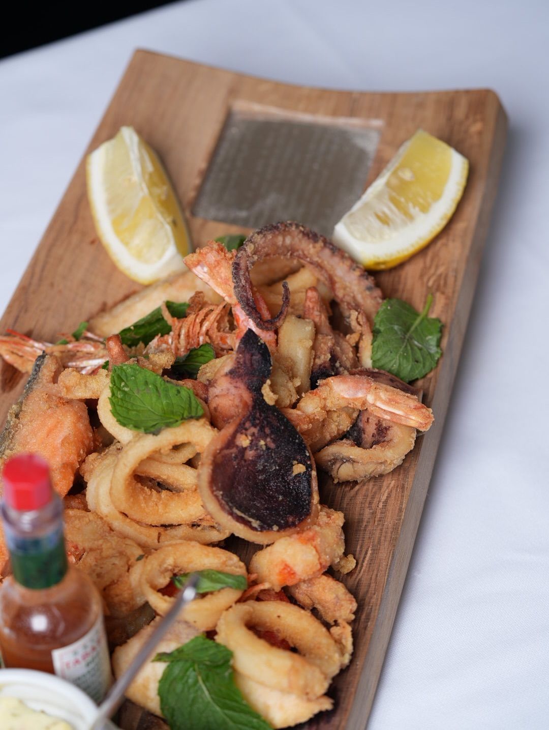 Pasta ai frutti di mare su un tagliere di legno con spicchi di limone, erbe aromatiche e bottiglia di salsa piccante.