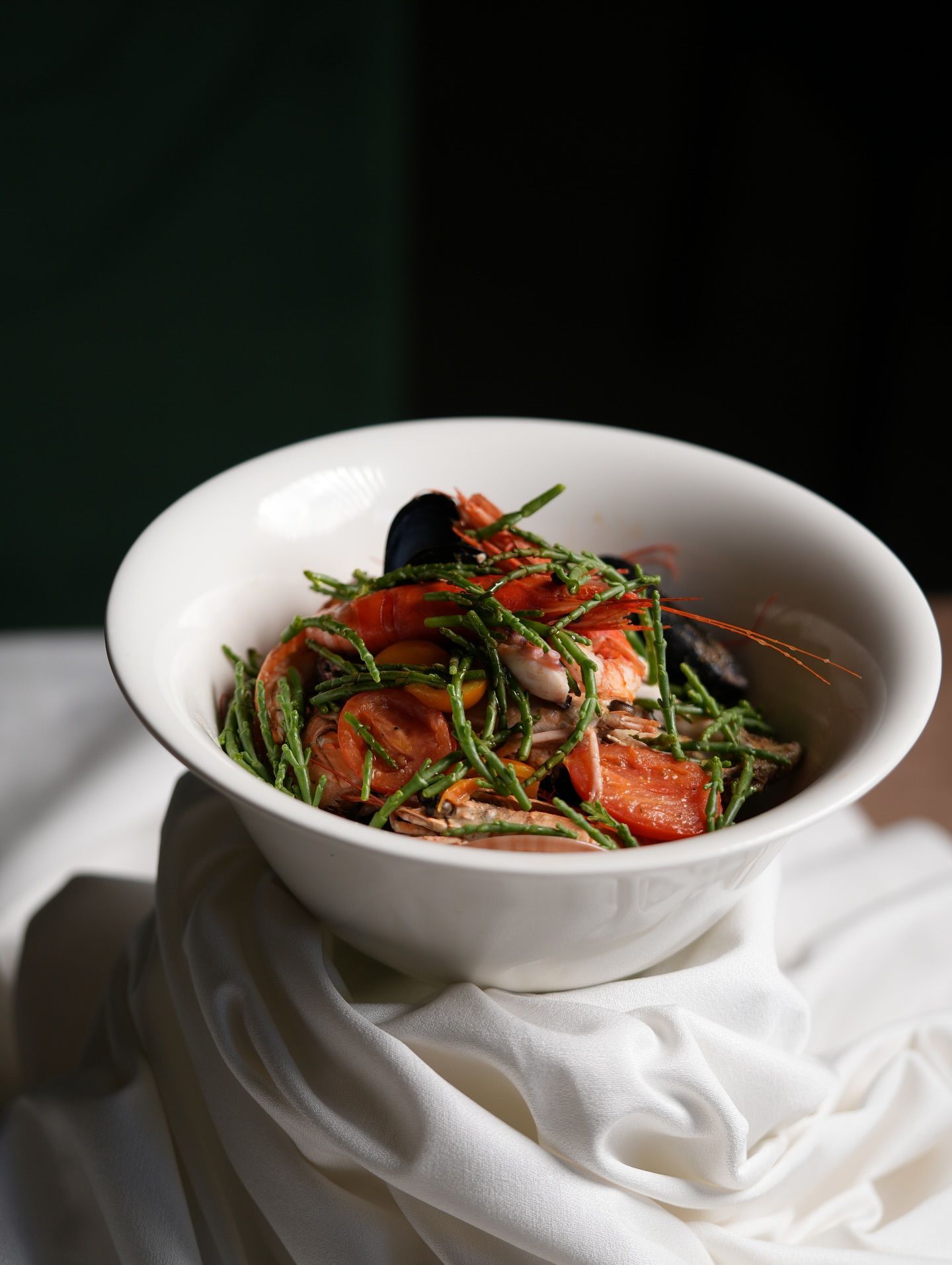 Ciotola bianca di pasta ai frutti di mare con salsa, guarnita con erbe aromatiche, adagiata su un drappeggio di stoffa bianca su uno sfondo scuro.