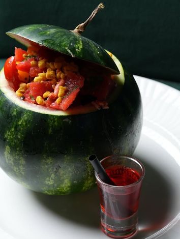 Anguria intagliata in una fruttiera piena di frutti di bosco, accanto a un bicchierino di succo rosso con cannuccia.