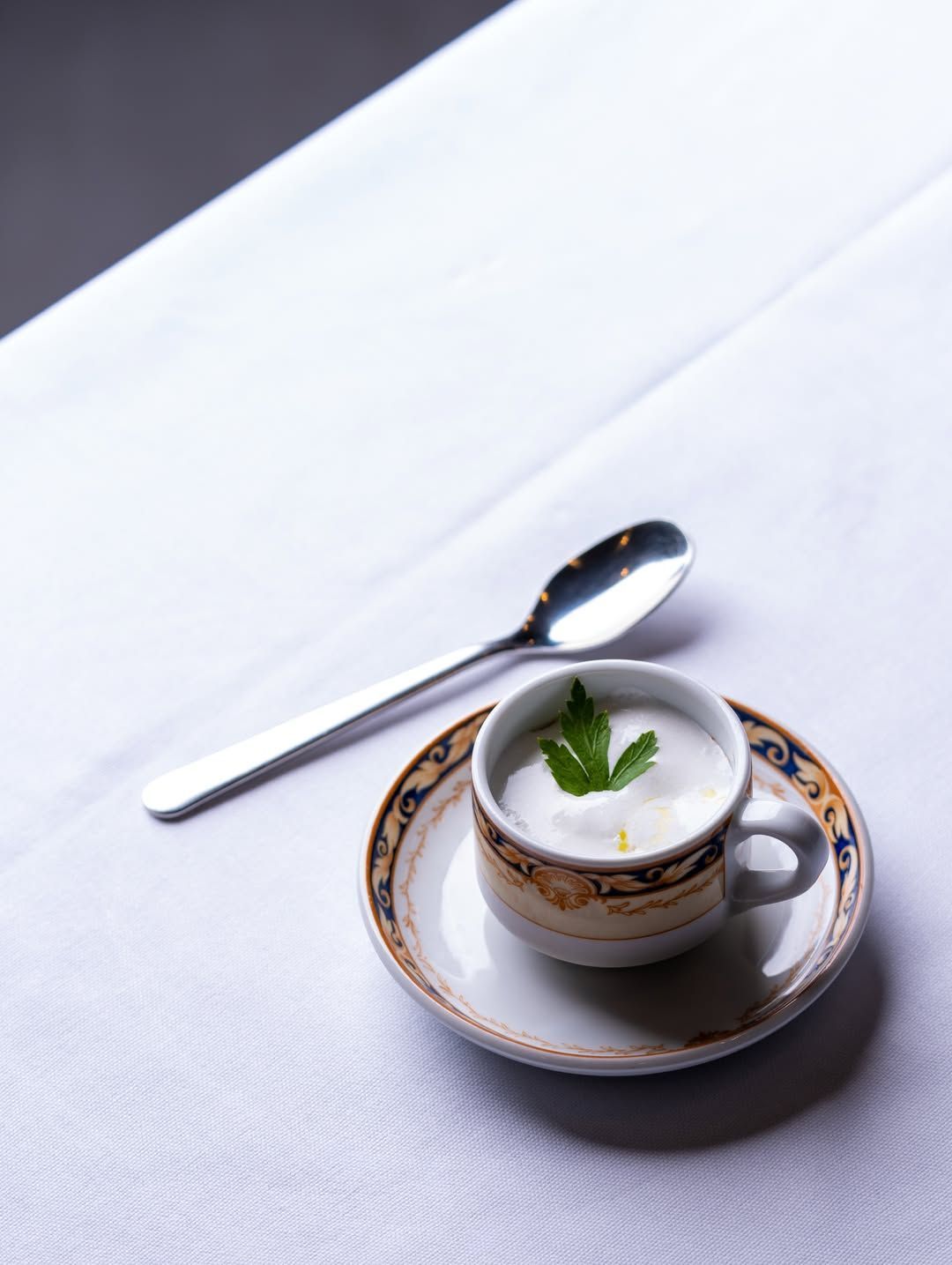 Un cappuccino bianco in una tazza decorata su un piattino, con un cucchiaino su una tovaglia bianca.