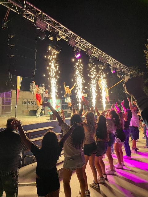 Persone che ballano a un concerto notturno all'aperto con luci brillanti sul palco e scintille simili a fuochi d'artificio.