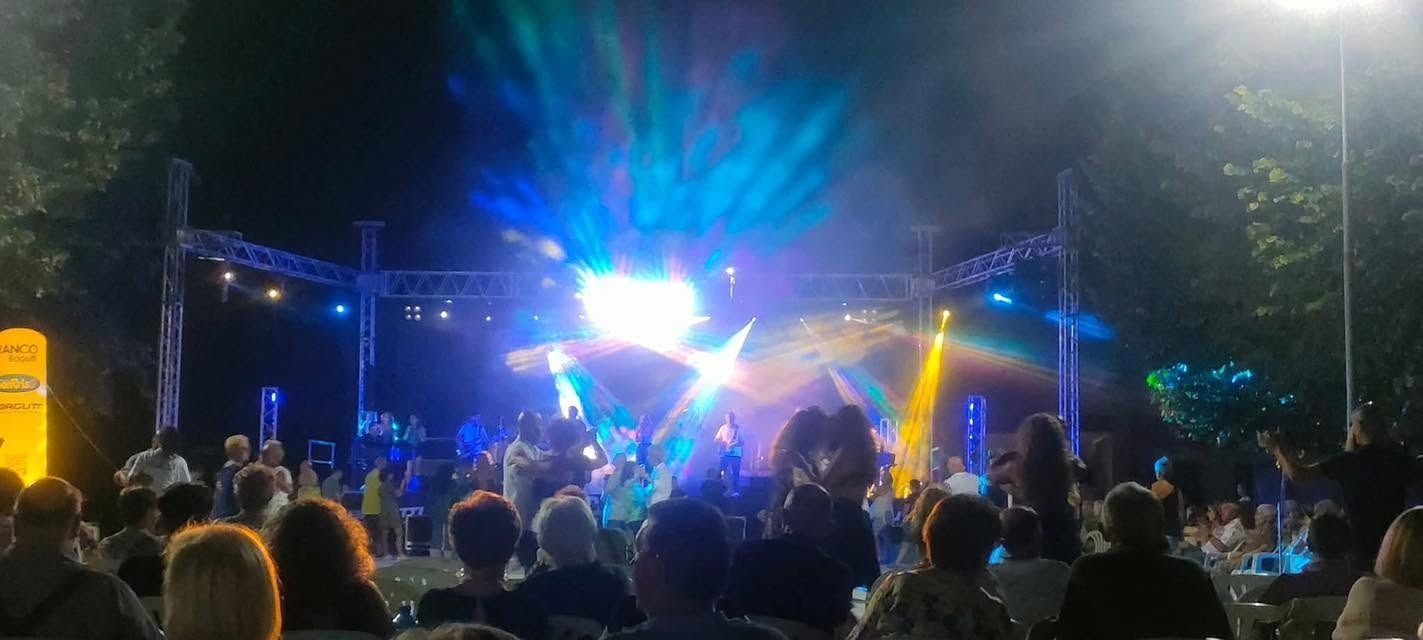 Pubblico che assiste a un concerto notturno all'aperto con luci di scena blu e gialle brillanti.