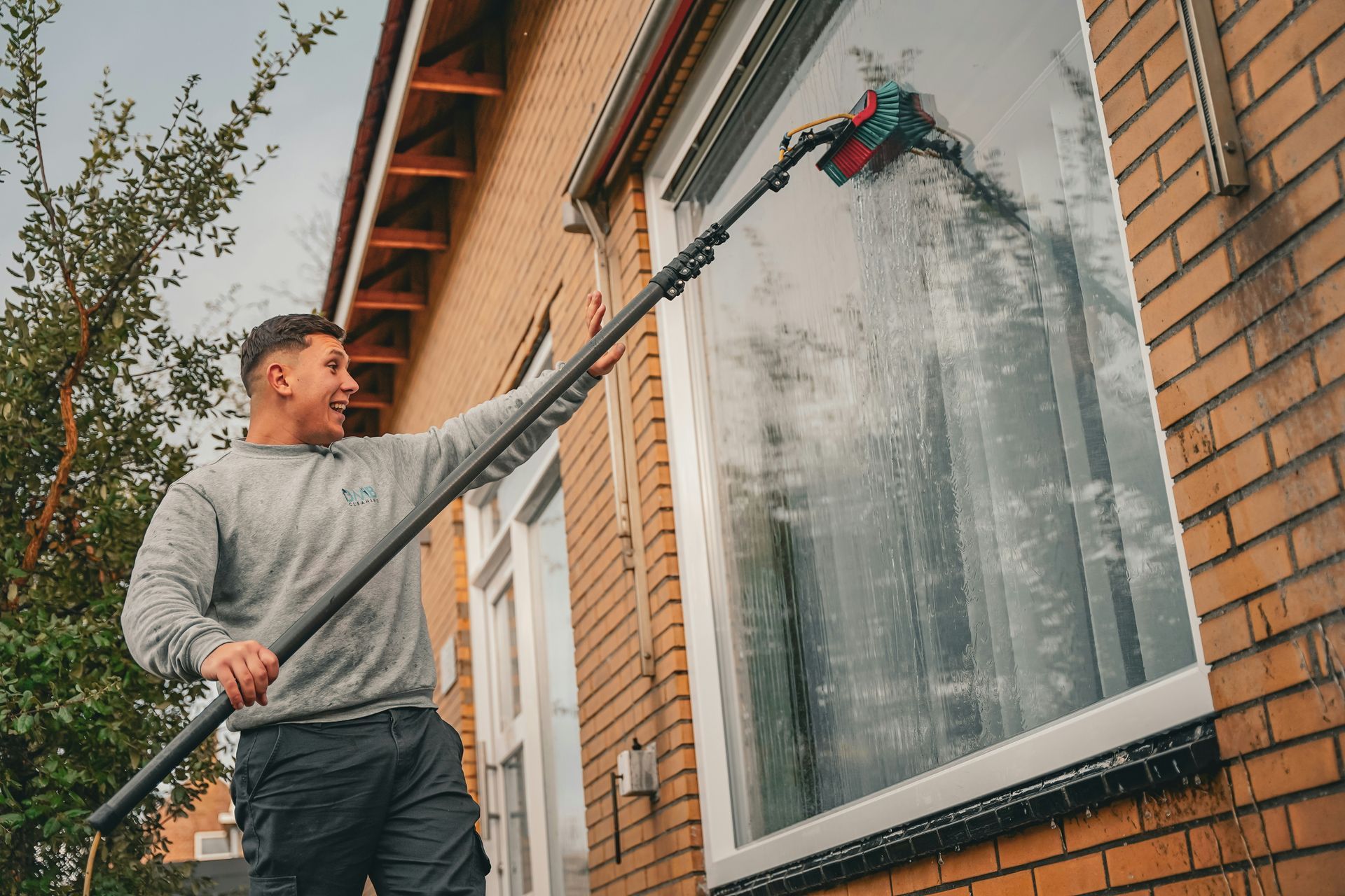 Man power-washes a brick house window with a long pole brush