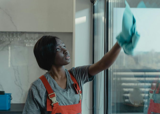 Person in orange overalls cleaning a window with a blue cloth in a kitchen.