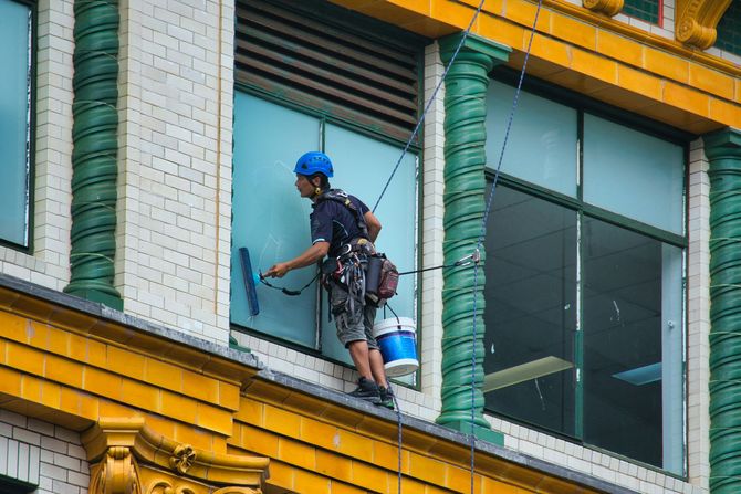 Worker suspended on a building facade painting a green window frame with a brush