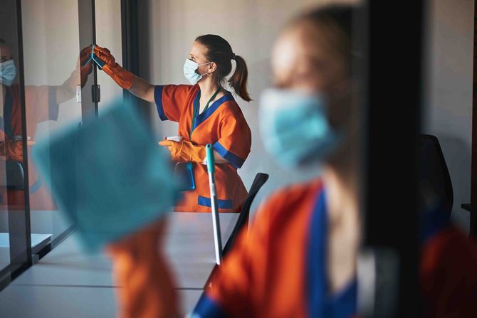 Workers in orange safety gear and masks disinfecting a glass wall in an office.