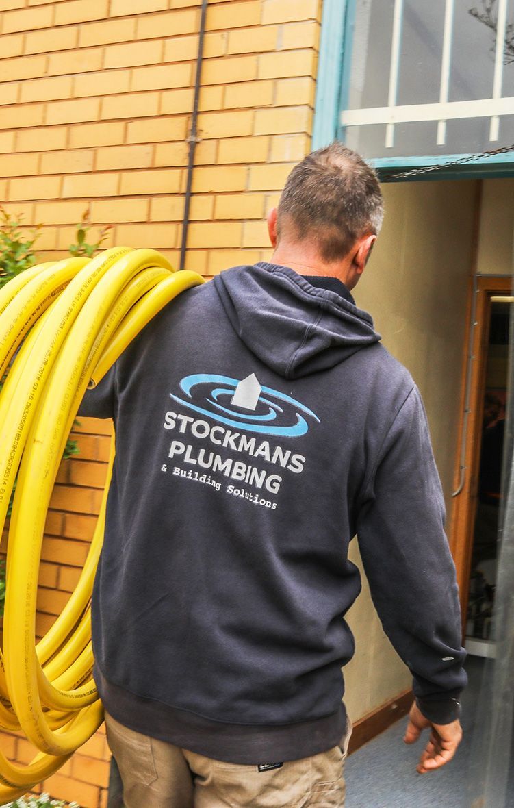 A man wearing a stockmans plumbing sweatshirt is carrying a yellow hose